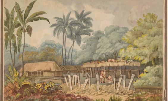 Two men are gathered under the shelter of a thatched roof structure. On top of the structure, offerings of meat and fish are strewn. In the foreground there are twelve signs or totems sticking out of the ground. To the left of the structure is a larger thatched roofed hut. The compound is surrounded by vegetation