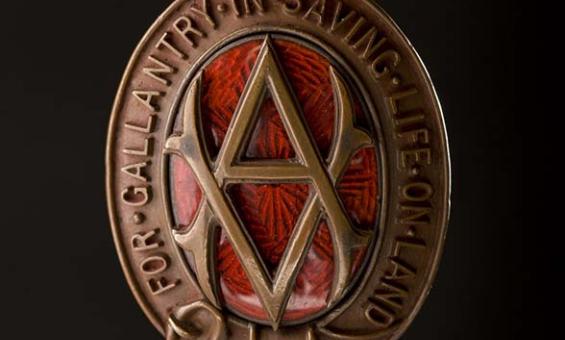 An oval shaped bronze medal. The oval is made of a belt buckled at the bottom. At the top is a stylised royal crown. In the centre of the oval is a monogram made up of an intertwined "V.A." Behind the monogram is pattered red glass. Around the oval in raised letters are the words 'For gallantry in saving life on land'. The medal is held by a ribbon of red and white vertical stripes