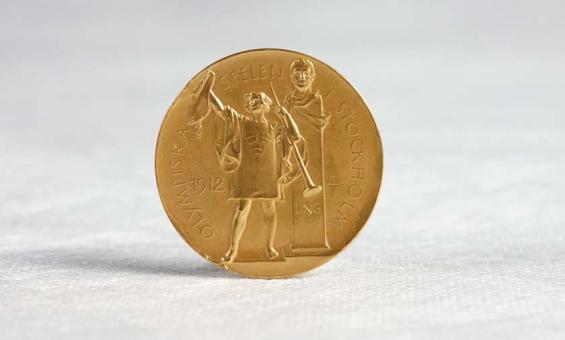A gold medal standing upright on a white cotton surface. The medal shows a figure holding a trumped and a wine amphorae. Behind the figure is a bust on a stand. In raised lettering around the edge of the medal are the words 'Olympiska Spelen Stockholm' The year '1912' is also present.