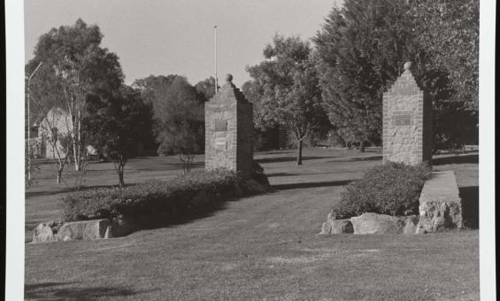A black and white photograph of the main garrison gates of the former Cowra Prisoner of War camp. The gates now sit in a grassy park surrounded by trees and bushes.