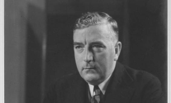 Australian Prime Minister Robert Menzies sits at a desk holding several pieces of paper. He is looking melancholic and stares off camera to the left. He is a middle-aged white man with blonde hair swept to one side. He wears a dark suit and tie with a dark tie. There is a black Bakelite telephone on the table in front of him