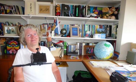 A photograph of a smiling woman seated in a wheelchair in front of a large bookcase and desk. She is wearing a white tshirt. The wheelchair has a control stick which sits just below her chin.