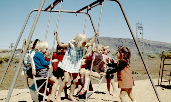 Photo of several young children in 1970s-era clothing playing on a swing set on a sunny cloudless day
