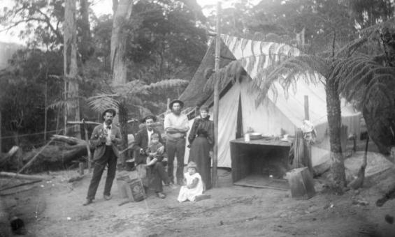 A black and white photograph of a family dressed in late 1800s style fashion. The are standing in a clearing in a forest and are standing in front of a tent.