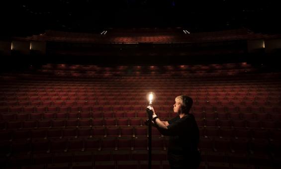 A person stands in an empty darkened theatre, illuminated by a single light bulb.