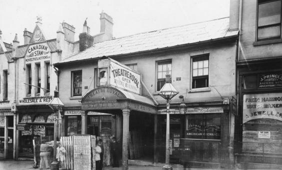 Black and white photograph of a row of shopfronts. One facade reads 'Theatre Royal: Open Every Evening'.