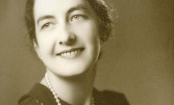 A sepia image of a woman in a black dress and pearl necklace, looking back and smiling over her shoulder.