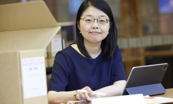 Woman with short dark hair wearing a navy short sleeve top smiling at the camera and sitting at a desk with a laptop, papers and a box