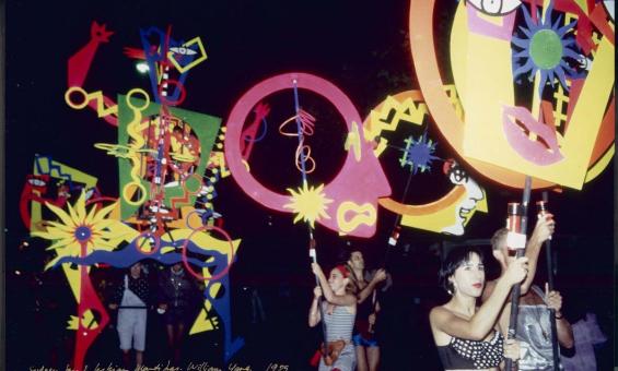 Participants in the 1995 Sydney Gay and Lesbian Mardi Gras parade carry large, colourful, abstract posters, flags and sculptures. The scene is set at night with vibrant shapes, patterns, and faces on display.