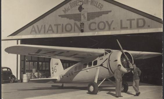 Two men standing and leaning against a small passenger plane in front of an aircraft hangar