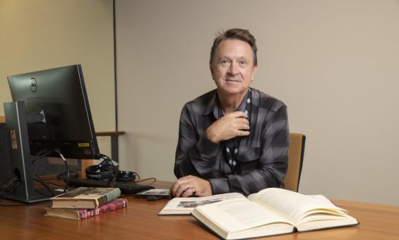 A man wearing a black and grey checkered shirt sitting at a desk with a computer and several books resting on it. He is leaning on the desk, facing forward and giving the camera a small smile