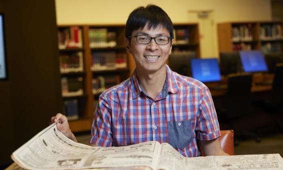 Man with short black hair and black glasses in a short-sleeved checkered shirt smiling and sitting at a desk with a Chinese language newspaper on it