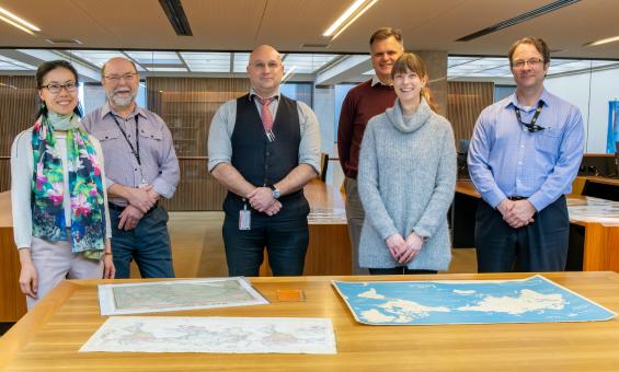 Two women and four men, who make up the Library's Special Collections Research & Support team, standing and smiling in the Special Collections Reading Room in front of a table with three maps on it