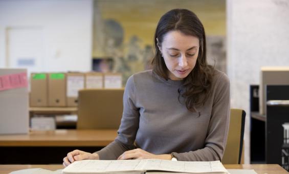 Woman with brown hair and eyes, wearing a brown sweater, sitting at a desk in the Special Collection Reading Room, looking down as she reads from an old book.