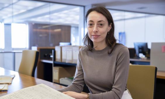 Woman with brown hair and eyes, wearing a brown sweater, sitting at a desk in the Special Collection Reading Room, smiling at the camera. On the desk in front of her is an old book of music.