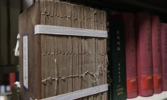 A large, old text with Chinese characters is bound with traditional thread stitching, sitting on a shelf, supported by a white fabric strap wrapped around its cover. The edges of the pages are uneven, showing wear with the stitching slightly frayed. Other colourful books with Chinese characters are lined up alongside it on the shelf.