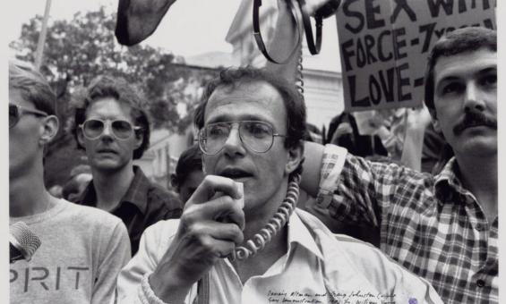 A black and white image of a group of mean demonstrating. The man front and centre is holding a microphone to his mouth, while the man to the right is holding a megaphone speaker up in the air. There are placards and flags in the air in the background.