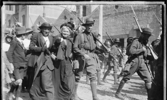 A black-and-white photograph shows a group of soldiers marching down a street with rifles over their shoulders. They are accompanied by civilians, who walk arm-in-arm with the soldiers, smiling and engaging with the crowd. The street is busy with people, and buildings can be seen in the background.