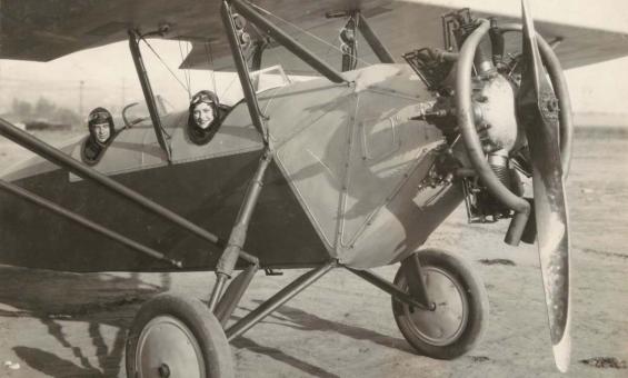 A black and white image of two people sitting in a 1920s style aeroplane while grounded.
