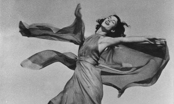 A black and white photograph of a woman in a dress twirling around with her eyes closed.