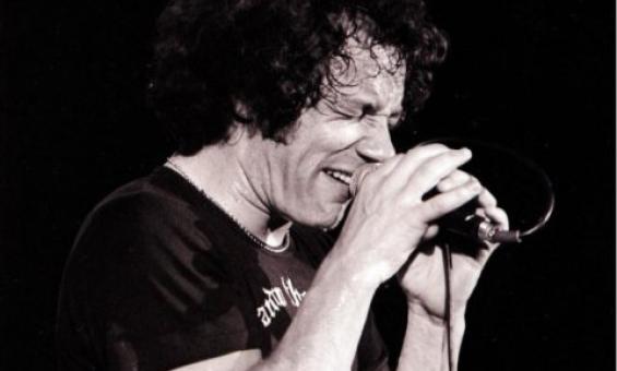 A black and white image of a man holding a microphone on a stand with 2 hands. He is holding the mic close to his mouth and has his eyes shut. He is wearing a black t-shirt and jeans.