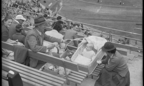 Family of three sitting in the crowded stands of a sports stadium while various events take place on the track and field. The parents seem to be enjoying a warm beverage while looking at their baby who lies in a stroller.