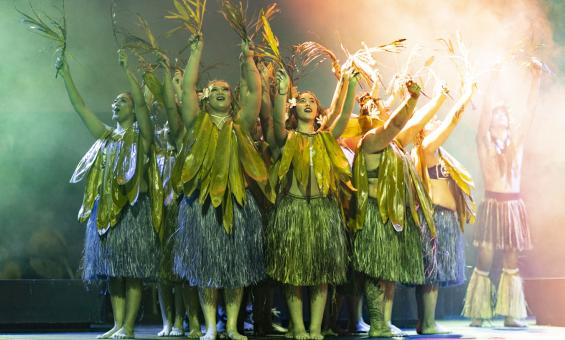 Group of dancers wearing large leaves around their necks and grass skirts, standing in a circle with their arms raised
