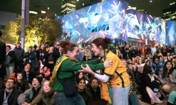 Two woman in green and gold clothing embracing excitedly in front of a crowd.