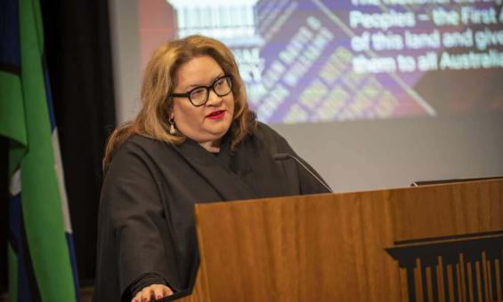 A person with long brown hair and glasses stands behind a wooden lectern, speaking into a microphone. The background features a screen displaying partially visible text and graphics. To the left, green and blue from the flag of the Torres Strait Islands is visible.