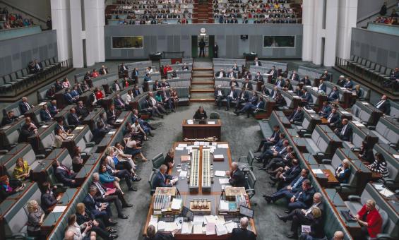 View of the Australian House of Representatives in Parliament House from slightly above. Members of the House of Representatives sit throughout the room, and members of the public sit in allocated areas