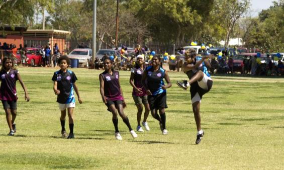 Six women smiling as they play football. One has just kicked the ball.