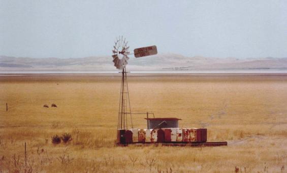 A windmill stands in a paddock surrounded by dry, yellow grass. Lake George in New South Wales is visible in the background.