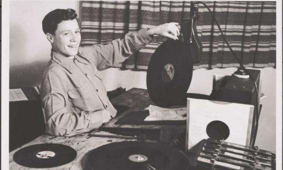 At the left of the photo, a young man with short dark hair is sitting down at a radio desk, smiling out at the camera and with his left hand he balances a record on its edge on the desk.