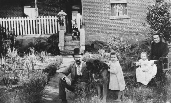 A black-and-white photo of a family posed outside a brick house with a white picket fence. In the foreground, a man in a bowler hat sits beside a large dog, while a young girl stands nearby. A seated woman holds a baby on her lap, and a child is visible on the steps of the house in the background. The garden surrounding them is filled with overgrown plants.