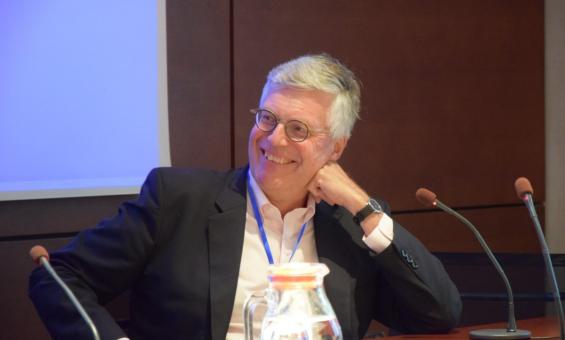 A man sitting at a table behind a microphone, turning his head and smiling.