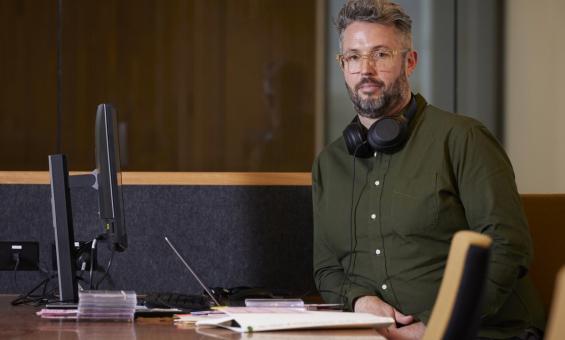 A man in glasses and a green shirt with headphones around his neck is sitting at a desk in front of a computer.