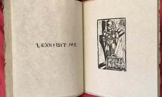 A large book sitting propped open on a red piece of material. The left hand page shows text that reads '1. Exhibit me' and the right hand page shows a stamped black ink image.