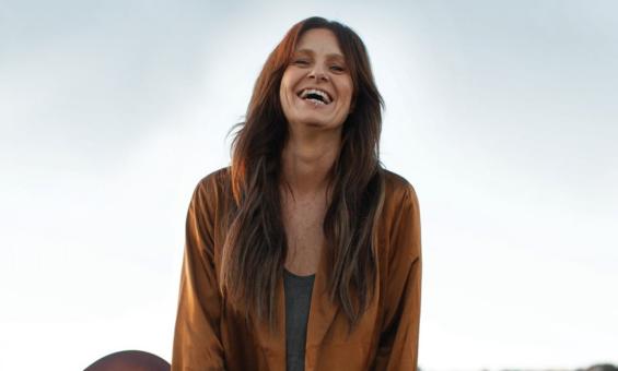 A woman with long brown hair, wearing a brown shirt and holding a guitar and smiling.