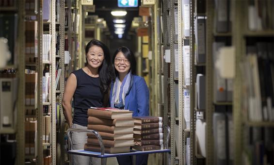 Two women standing behind a trolley with books on it in the Library stacks.