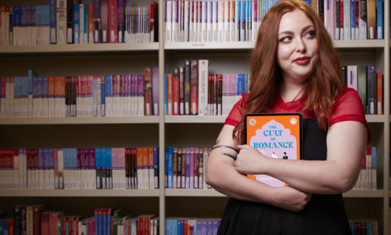 Woman with red hair standing in front of a bookshelf of romance books, holding a tablet with the cover of a romance book on the screen. She is facing forward with her back to the bookshelf, looking to the side and slightly smiling