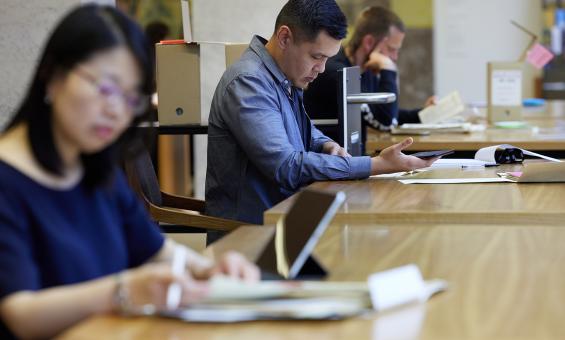 Three people sitting at wooden tables, looking at National Library collection material.