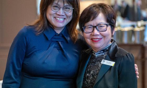 Two people standing next to each other, smiling. One has shoulder-length dark brown hair and wears glasses and a dark blue dress with a bow detail at the neckline. The other has short dark hair, also wearing glasses, and a black jacket with a floral pattern on the lapel, along with a name tag.