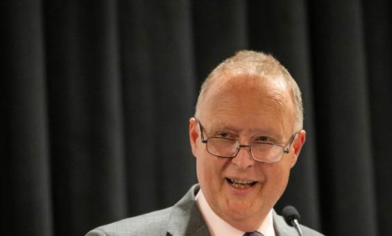 Rupert Myer AO, wearing a grey suit with a pink shirt and patterned tie, speaks at a podium with a microphone. He is smiling warmly and wearing glasses, addressing an audience against a backdrop of dark curtains.