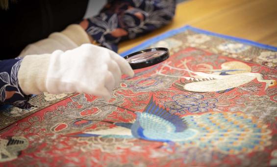 A person wearing white gloves examining a colorful peacock embroidery with a magnifying glass, focusing on the intricate details of the artwork.