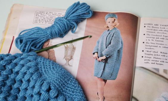 An old magazine showing a vintage crochet pattern. Laying on top of the magazine is a blue crochet item in progress.