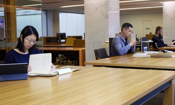 Three people, one woman and two men working at a long table. They are each doing their own research using laptops, notebooks and other resources