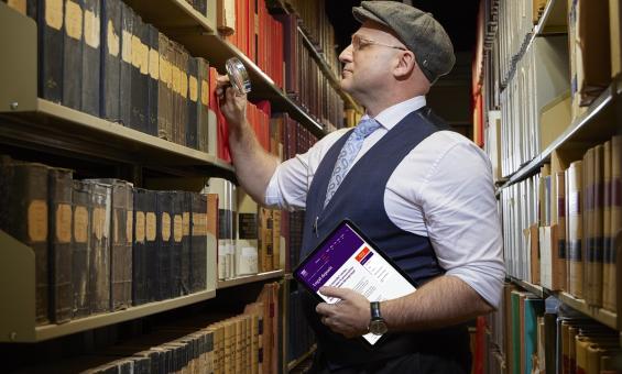 A person standing in front of a bookshelf, holding a magnifying glass up to the shelf. They are also holding an ipad with a website showing on it.