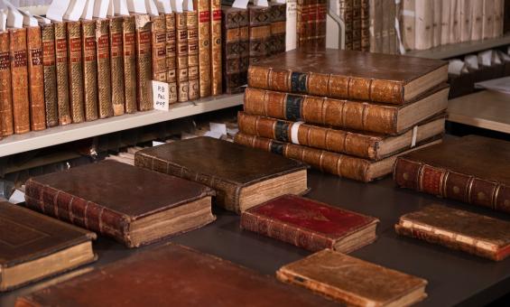Shelf and table with dozens of old, leather-bound books, some with gold designs or writing on the spines
