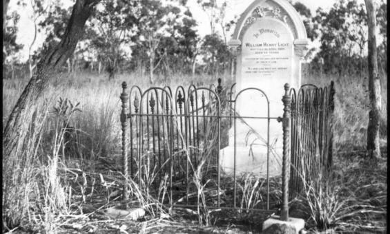 Black and white photograph of a gravestone surrounded by a wrought iron fence. The grave is situated in a grassy, rural setting with a few trees scattered around. The gravestone has an arched top and an inscription in memory of William Henry Light.