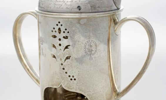 A silver polished kettle with ornate cutouts and inscription and printed crest. It has a tealight underneath.